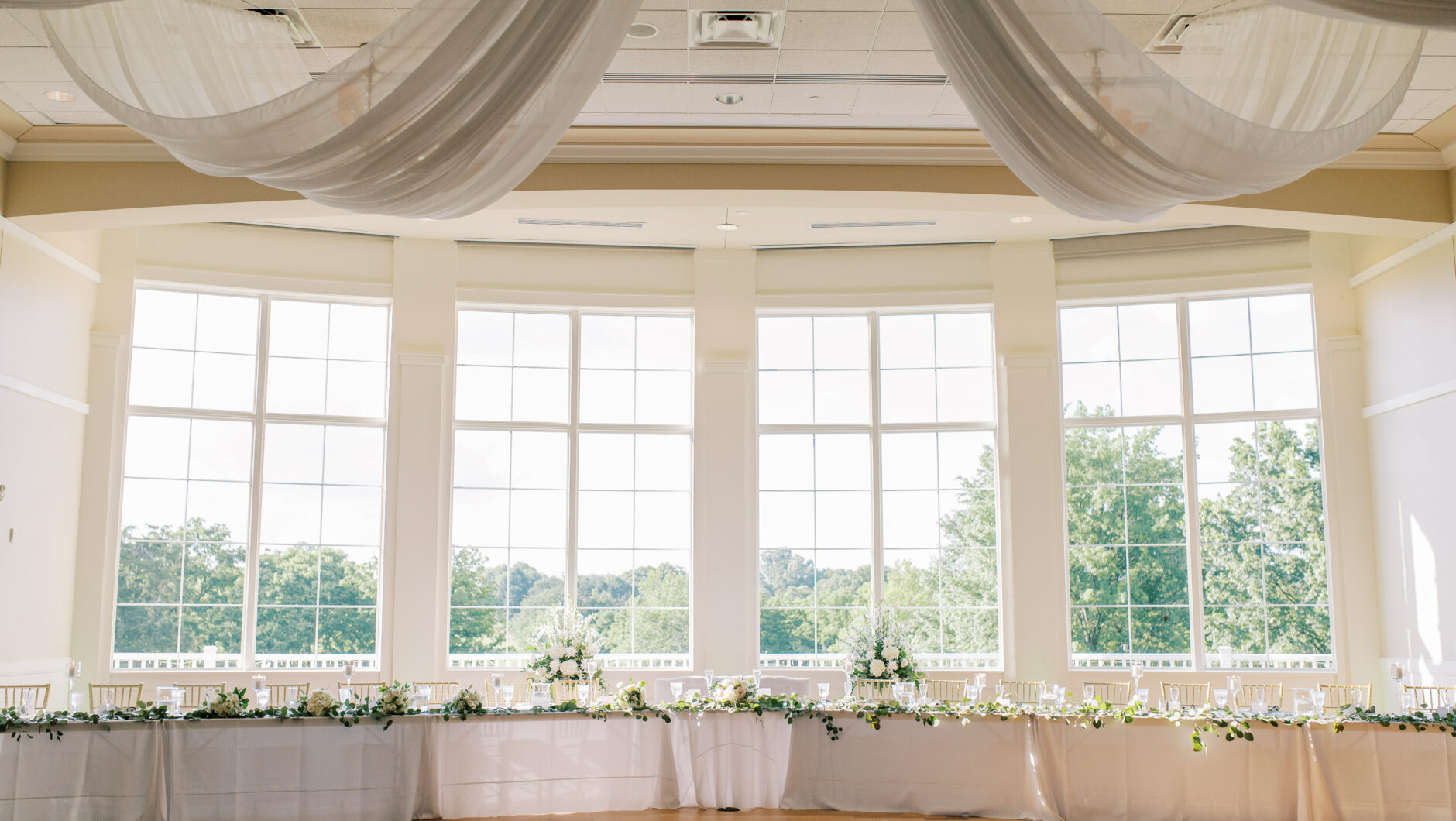 Open room with floor to ceiling windows, long white table with gold chairs and flowers, and big white draping curtains hanging from the ceiling