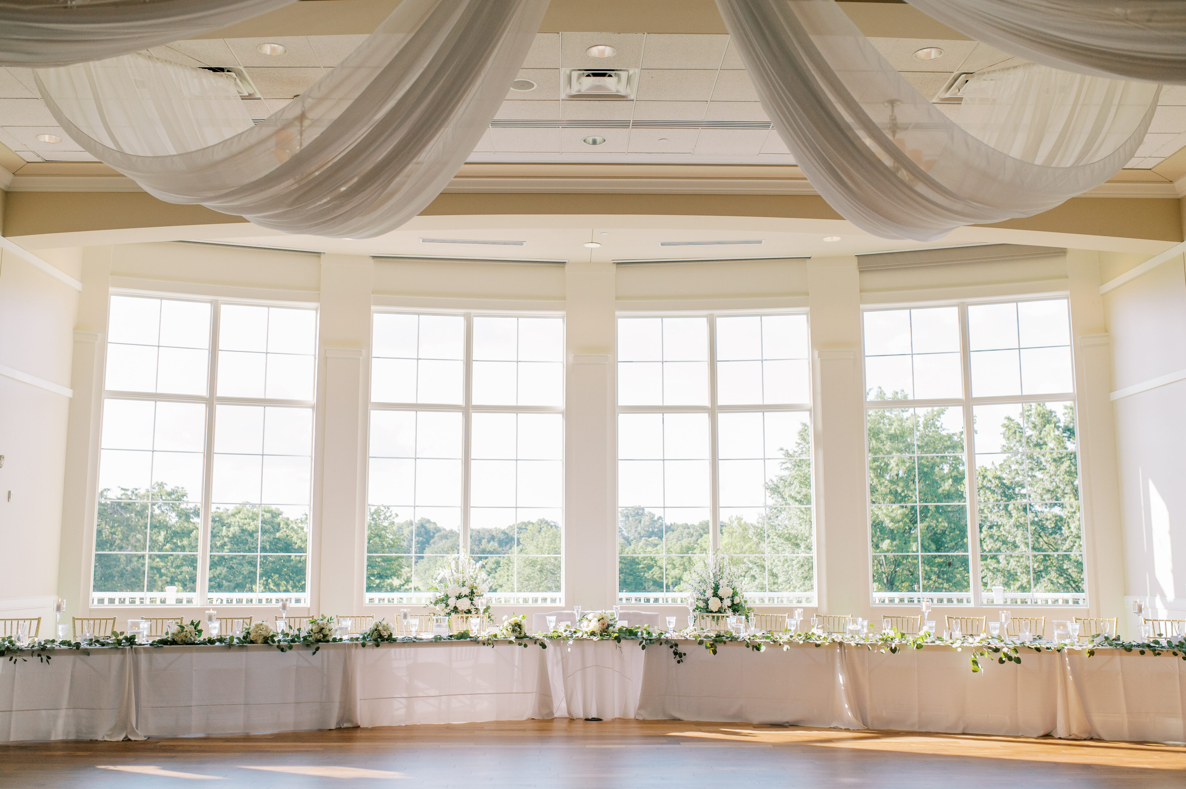 Open room with floor to ceiling windows, long white table with gold chairs and flowers, and big white draping curtains hanging from the ceiling
