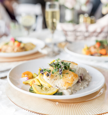 A plated meal of grilled fish with mashed potatoes, sautéed vegetables, and microgreens, with champagne in background.