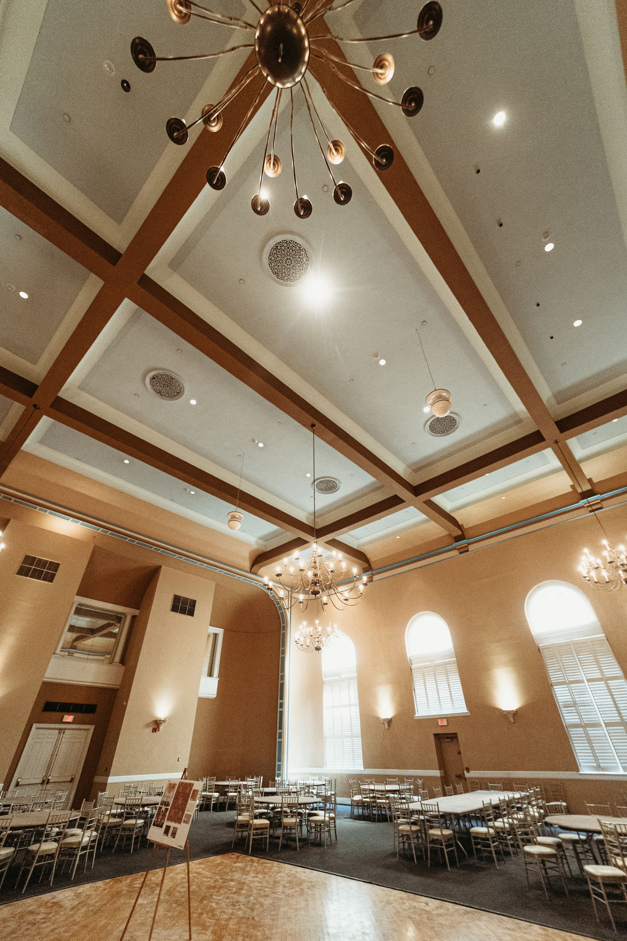 Ballroom with round tables, chairs, chandeliers, and high ceilings, set up for an event or reception.