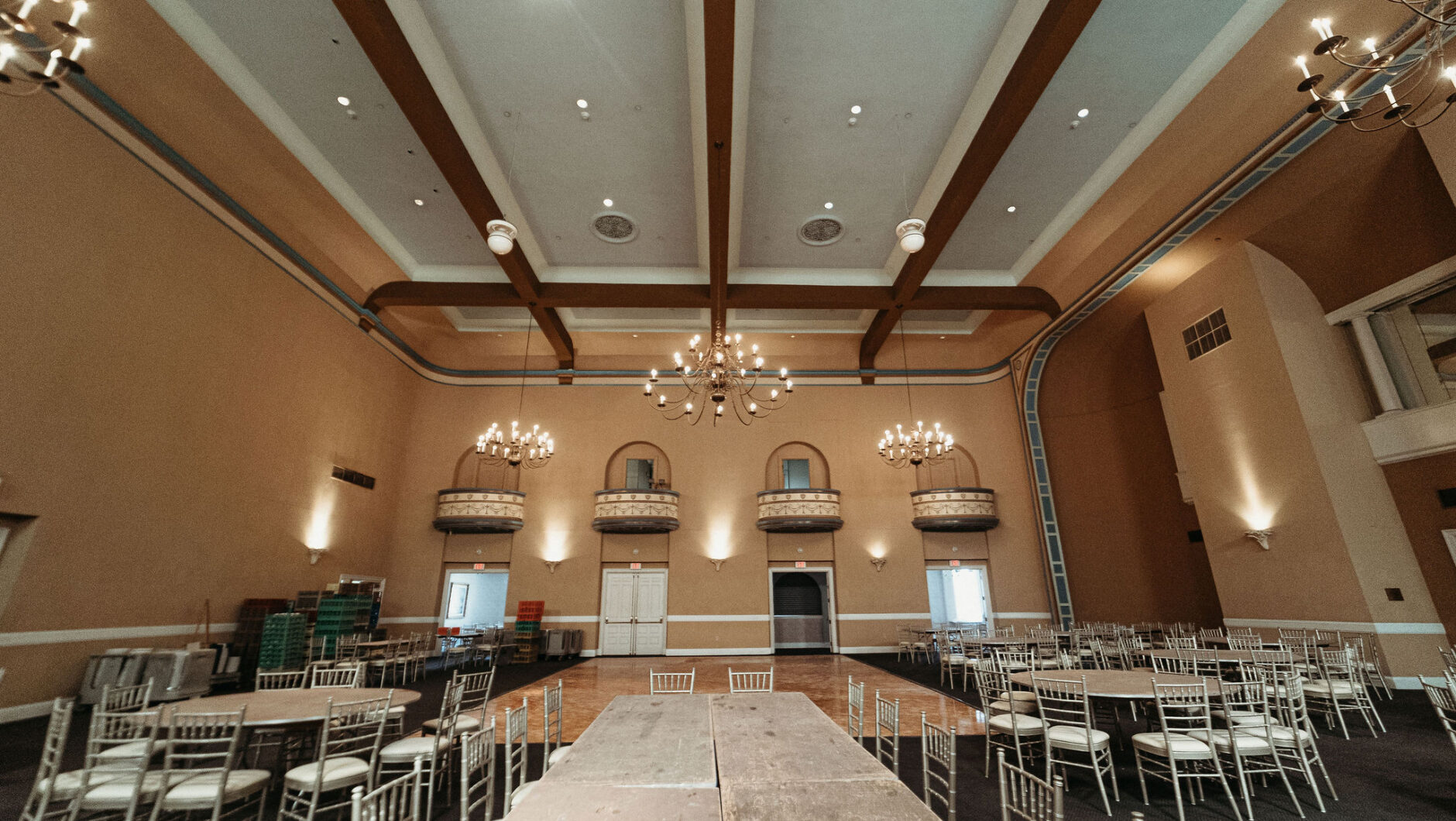 Large empty banquet hall with chandeliers, round tables, and stacked chairs against peach-colored walls.