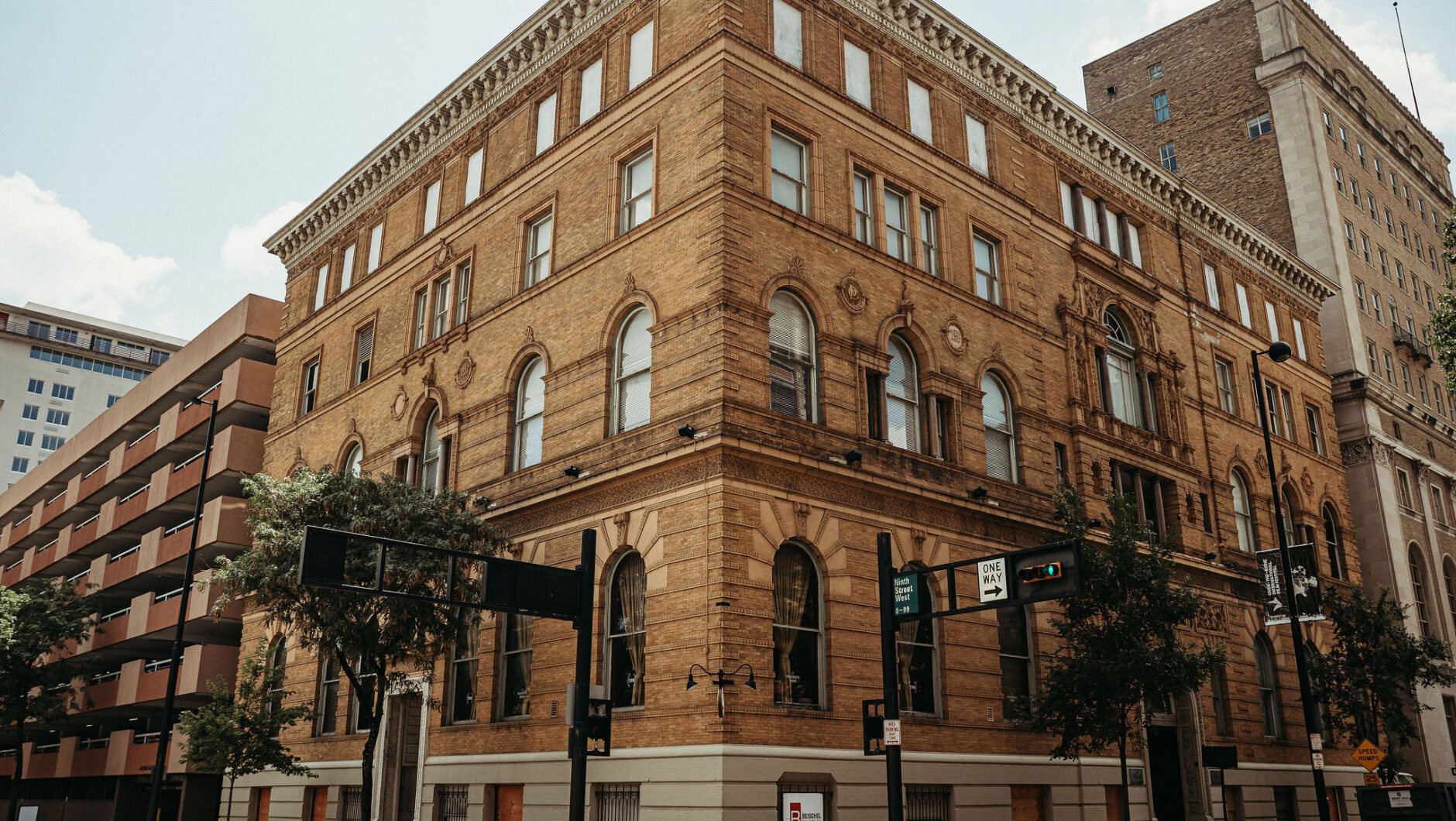 A historic, four-story brick building stands at a street corner with large windows and trees nearby.
