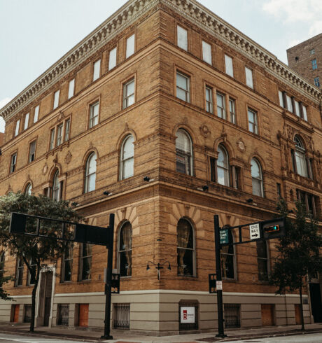 A historic, four-story brick building stands at a street corner with large windows and trees nearby.