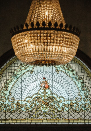 A crystal chandelier hangs in front of a stained glass window with an ornate floral design.