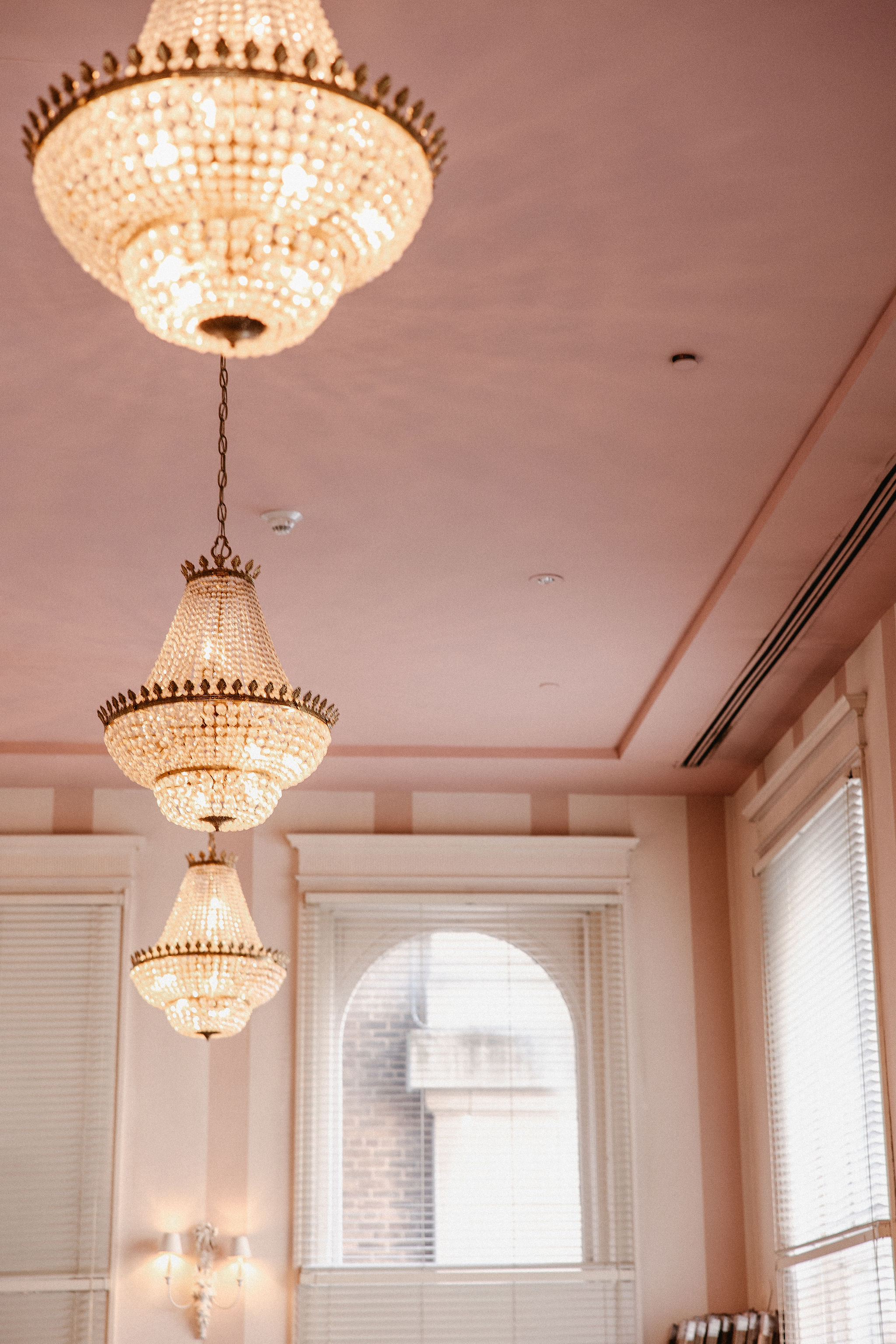 Three ornate chandeliers hang from a pink ceiling in an elegant room with large windows and white blinds.
